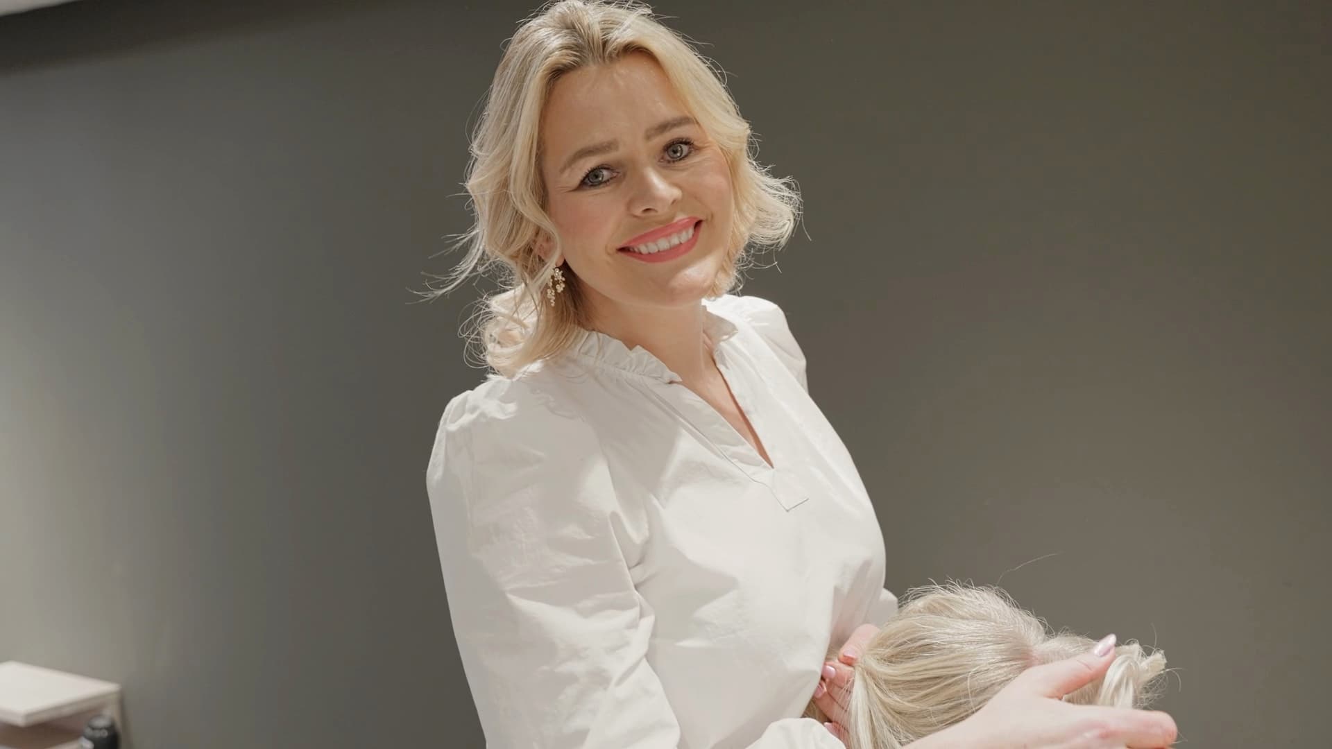 Frisør i hvit bluse smiler mens hun jobber med håret til en kunde